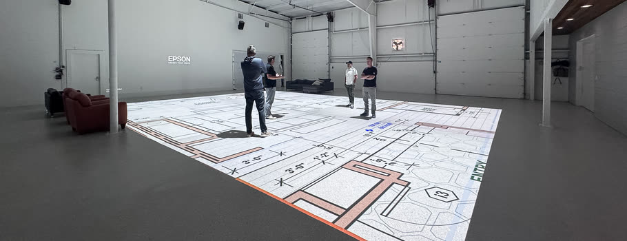 Four people spread across a floor plan projected at full scale on the studio floor, looking at the rooms around them.