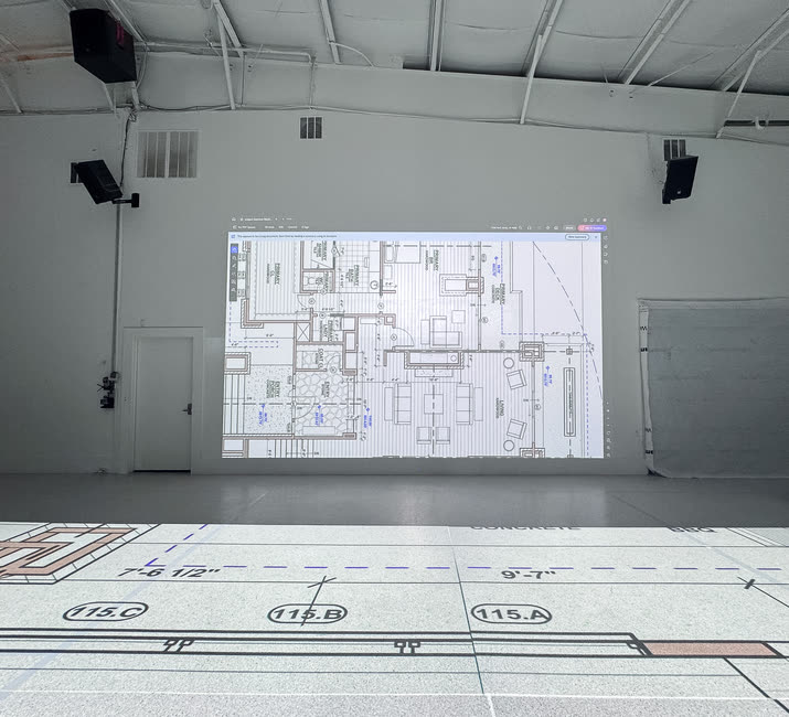 A floor plan on the studio wall display, with the same plan projected at full scale on the floor in front of it, door tags and dimensions readable.