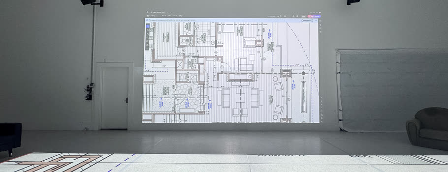 A floor plan on the studio wall display, with the same plan projected at full scale on the floor in front of it, door tags and dimensions readable.