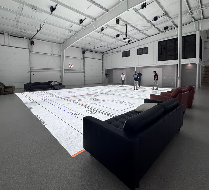 The studio floor with a plan projected across it, sofas and armchairs set at its edge, and four people standing at its far end.