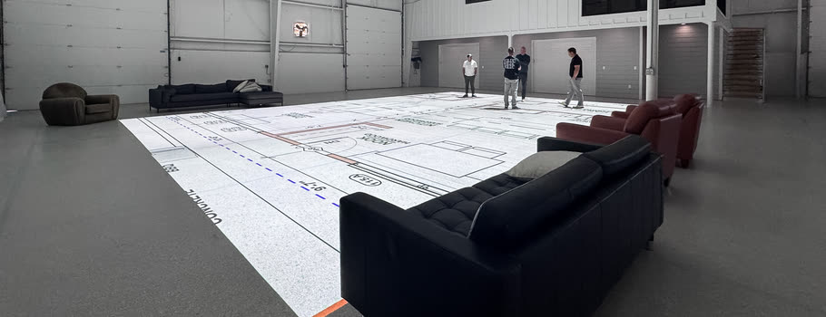The studio floor with a plan projected across it, sofas and armchairs set at its edge, and four people standing at its far end.