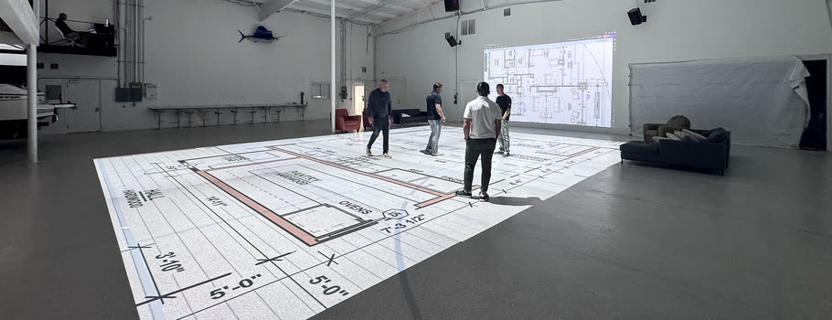 Four people walking a floor plan projected at full scale, with the same drawing displayed on the wall behind them.