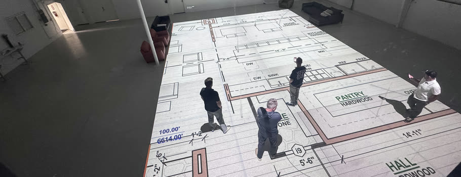 A floor plan projected at full scale across the studio floor, with four people standing inside its rooms. Dimensions and room labels — PANTRY, HALL — are readable underfoot.